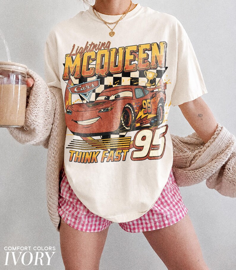 K&ouml;nnte beinhalten: Elfenbeinfarbenes T-Shirt mit Lightning McQueen aus dem Film Cars. Die Grafik zeigt das Auto, die Nummer 95 und den Schriftzug "Think Fast". Das Shirt wird mit einer rosa-wei&szlig; karierten Shorts kombiniert.