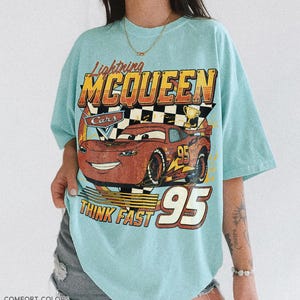 K&ouml;nnte beinhalten: Hellblaues T-Shirt mit einer Grafik von Lightning McQueen aus dem Film Cars. Das Design enth&auml;lt den Text "Lightning McQueen", "Cars", "Think Fast" und die Nummer "95". Das Shirt ist von der Marke Comfort Colors in der Farbe Charky Mint.