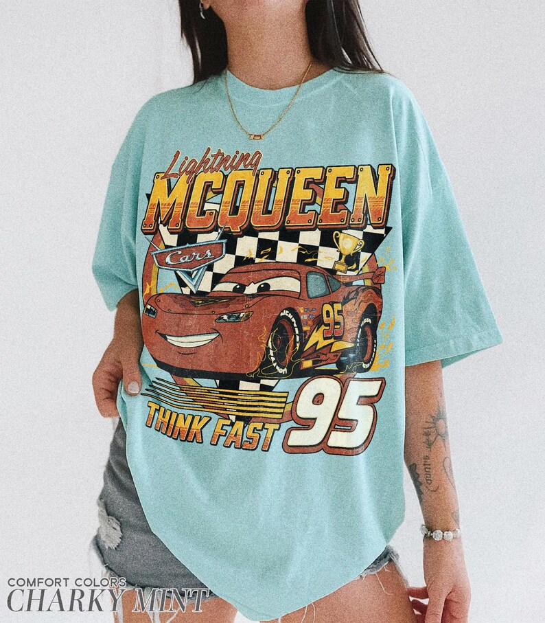 K&ouml;nnte beinhalten: Hellblaues T-Shirt mit einer Grafik von Lightning McQueen aus dem Film Cars. Das Design enth&auml;lt den Text "Lightning McQueen", "Cars", "Think Fast" und die Nummer "95". Das Shirt ist von der Marke Comfort Colors in der Farbe Charky Mint.