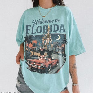 May include: A light blue t-shirt with the words "Welcome to Florida" and a graphic of Mickey and Minnie Mouse in a red convertible in front of a castle. The shirt also has the text "EST. 1971".