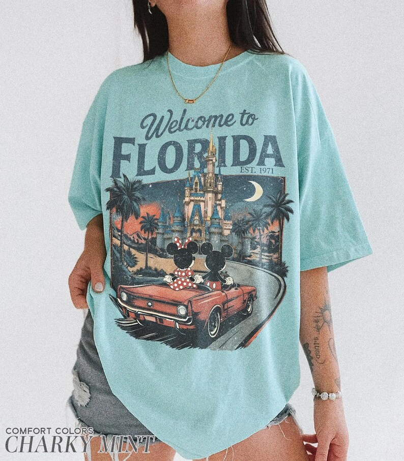 May include: A light blue t-shirt with the words "Welcome to Florida" and a graphic of Mickey and Minnie Mouse in a red convertible in front of a castle. The shirt also has the text "EST. 1971".