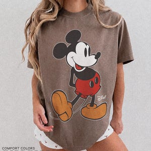 K&ouml;nnte beinhalten: Espressofarbenes T-Shirt mit einer gro&szlig;en Grafik von Mickey Mouse in seiner klassischen Pose. Die Zeichentrickfigur ist in Schwarz, Wei&szlig;, Rot und Beige dargestellt. Auf dem Shirt stehen die Worte "Comfort Colors" und "Espresso".