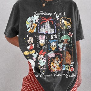 May include: Dark gray t-shirt featuring a colorful graphic of Disney characters and landmarks. The design includes Mickey Mouse, Donald Duck, and Cinderella's castle. Text reads "Walt Disney World" and "Most Magical Place on Earth 2026".