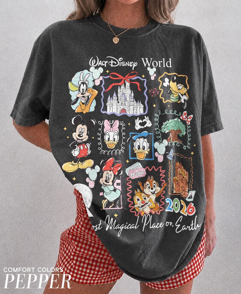 May include: Dark gray t-shirt featuring a colorful graphic of Disney characters and landmarks. The design includes Mickey Mouse, Donald Duck, and Cinderella's castle. Text reads "Walt Disney World" and "Most Magical Place on Earth 2026".