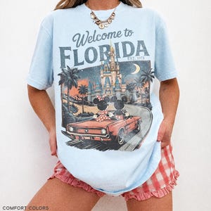 May include: Light blue t-shirt with a graphic that says "Welcome to Florida" and features a vintage illustration of Mickey and Minnie Mouse in a car, with a castle and palm trees. The shirt is made of chambray fabric.