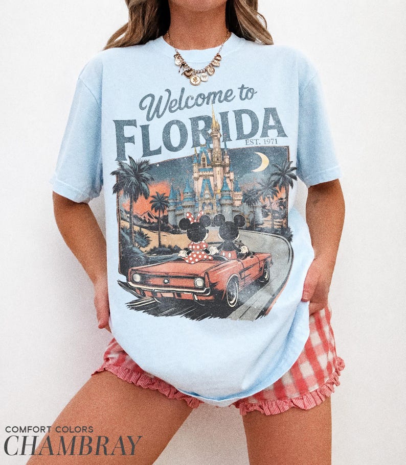 May include: Light blue t-shirt with a graphic that says "Welcome to Florida" and features a vintage illustration of Mickey and Minnie Mouse in a car, with a castle and palm trees. The shirt is made of chambray fabric.