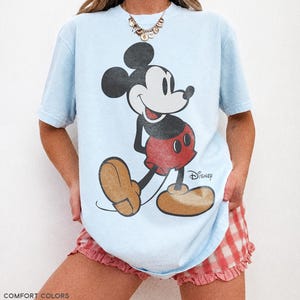 K&ouml;nnte beinhalten: Hellblaues T-Shirt mit einem Vintage-Mickey-Mouse-Design. Die Cartoonfigur ist in Schwarz, Rot und Beige gehalten. Auf dem T-Shirt steht unten "COMFORT COLORS CHAMBRAY". Ein klassisches Kleidungsst&uuml;ck im Disney-Stil.