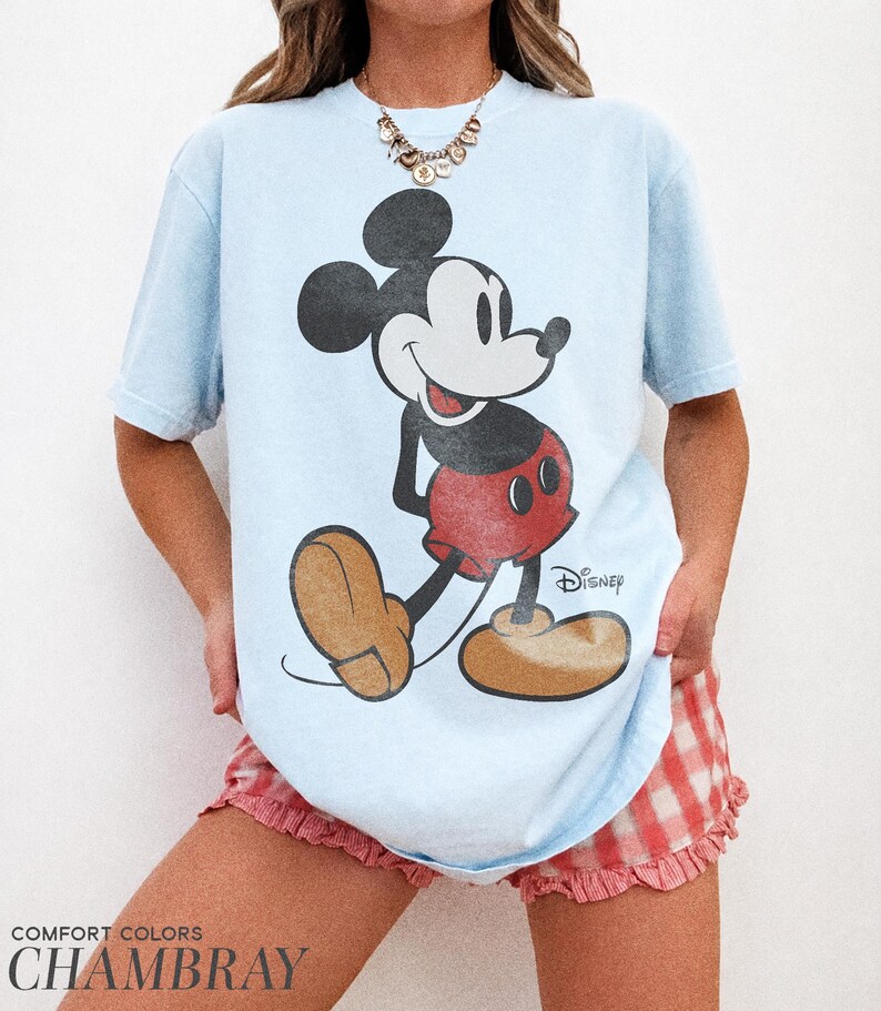 K&ouml;nnte beinhalten: Hellblaues T-Shirt mit einem Vintage-Mickey-Mouse-Design. Die Cartoonfigur ist in Schwarz, Rot und Beige gehalten. Auf dem T-Shirt steht unten "COMFORT COLORS CHAMBRAY". Ein klassisches Kleidungsst&uuml;ck im Disney-Stil.