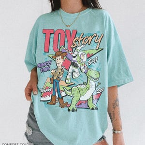 May include: A light blue t-shirt featuring the text "TOY STORY" in red and a graphic of Woody, Buzz Lightyear, and Rex. The shirt also includes the text "SINCE 1995" and "COMFORT COLORS CHARKY MINT".