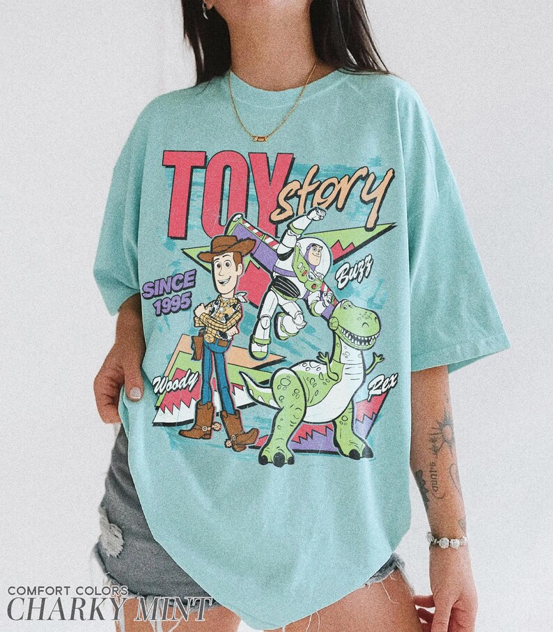 May include: A light blue t-shirt featuring the text "TOY STORY" in red and a graphic of Woody, Buzz Lightyear, and Rex. The shirt also includes the text "SINCE 1995" and "COMFORT COLORS CHARKY MINT".