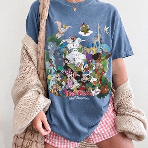Peut inclure: T-shirt bleu jean avec un motif color&eacute; de personnages Disney et le texte "Magic Kingdom Walt Disney World". Le t-shirt porte &eacute;galement le texte "Comfort Colors Blue Jean" en bas.