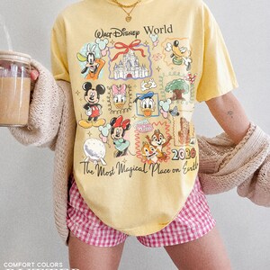 May include: A yellow t-shirt with a Walt Disney World graphic featuring various cartoon characters and the text "The Most Magical Place on Earth". The shirt also has the year "2026" printed on it. The model is wearing pink and white checkered shorts.