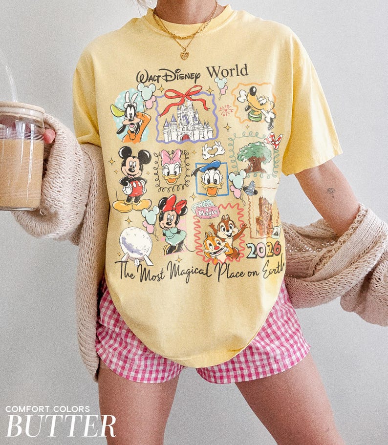 May include: A yellow t-shirt with a Walt Disney World graphic featuring various cartoon characters and the text "The Most Magical Place on Earth". The shirt also has the year "2026" printed on it. The model is wearing pink and white checkered shorts.