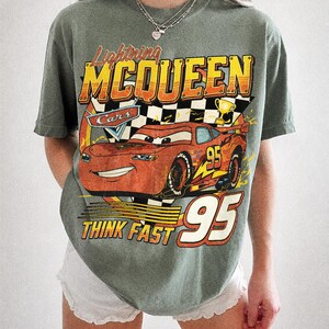 K&ouml;nnte beinhalten: Moosgr&uuml;nes T-Shirt mit einem Aufdruck von Lightning McQueen aus dem Film Cars. Das Design enth&auml;lt den Text "Lightning McQueen", "Cars", "Think Fast" und die Nummer "95". Das Shirt ist von der Marke Comfort Colors.