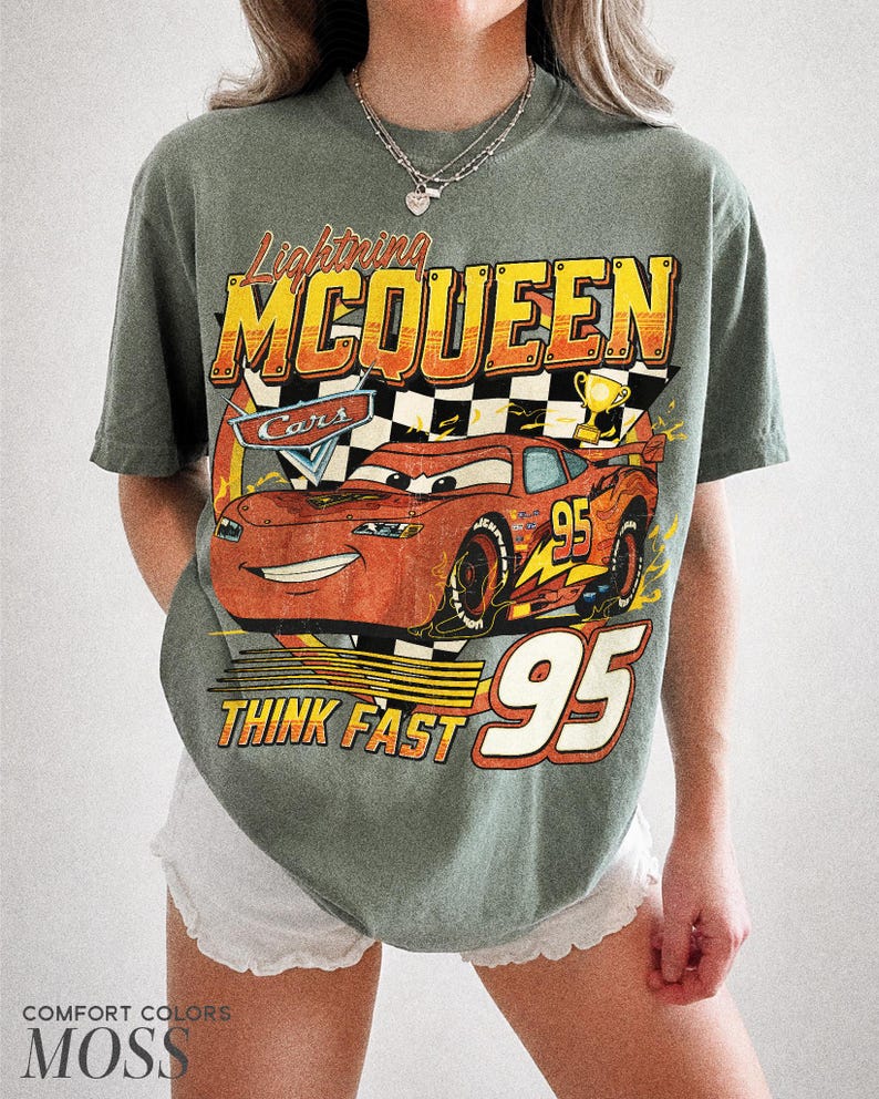 K&ouml;nnte beinhalten: Moosgr&uuml;nes T-Shirt mit einem Aufdruck von Lightning McQueen aus dem Film Cars. Das Design enth&auml;lt den Text "Lightning McQueen", "Cars", "Think Fast" und die Nummer "95". Das Shirt ist von der Marke Comfort Colors.