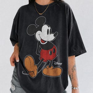 K&ouml;nnte beinhalten: Schwarzes T-Shirt mit einem klassischen Mickey Mouse-Motiv. Das Design zeigt Mickeys Gesicht, rote Shorts und braune Schuhe. Das Wort "Disney" ist darunter gedruckt. Das T-Shirt ist mit "Comfort Colors Black" gekennzeichnet.