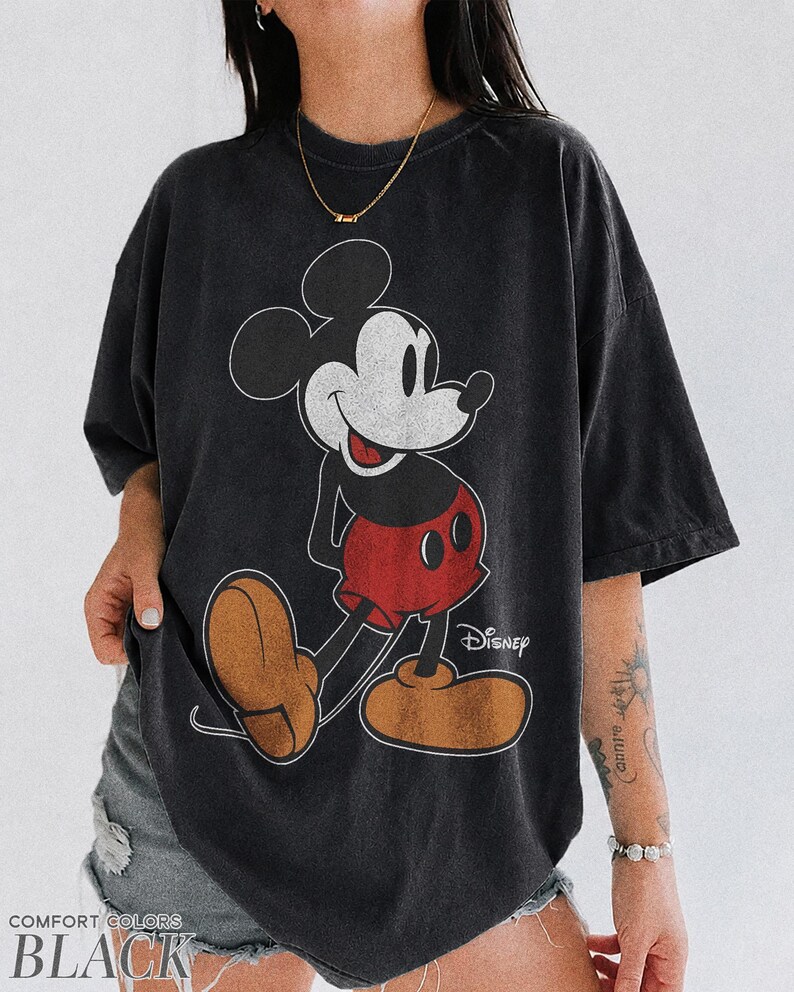 K&ouml;nnte beinhalten: Schwarzes T-Shirt mit einem klassischen Mickey Mouse-Motiv. Das Design zeigt Mickeys Gesicht, rote Shorts und braune Schuhe. Das Wort "Disney" ist darunter gedruckt. Das T-Shirt ist mit "Comfort Colors Black" gekennzeichnet.