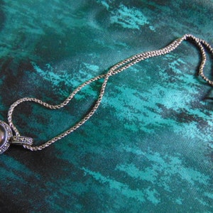 May include: A silver necklace with a round pendant featuring a dark gray stone surrounded by smaller stones. The necklace is on a teal fabric background.