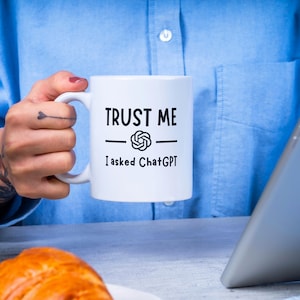 Op de afbeelding: Witte keramische mok met de tekst "TRUST ME" boven een afbeelding en de tekst "I asked ChatGPT" in het zwart. De mok wordt vastgehouden door een persoon die een blauw overhemd draagt. Op de voorgrond is een croissant te zien.