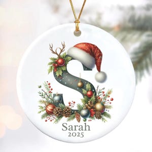 May include: A white Christmas ornament with a gold string. The ornament features a festive design with the letter "S" decorated with holly, berries, and a Santa hat. The name "Sarah" and the year "2025" are printed below.