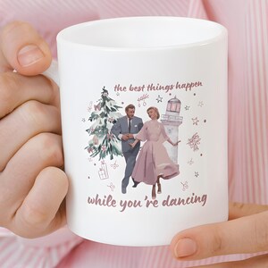 May include: White ceramic mug featuring a vintage illustration of a couple dancing near a Christmas tree and a lighthouse. The mug has the text "the best things happen while you're dancing" in a mauve color.