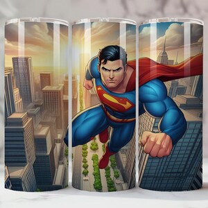 May include: Three stainless steel tumblers with a Superman design. The artwork shows Superman flying over a city, wearing a red cape and blue suit. The background features a sunset over the city.