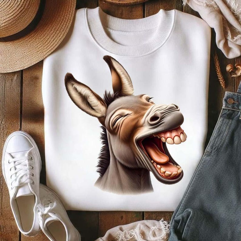Op de afbeelding: Witte crewneck sweatshirt met een gedetailleerde illustratie van een lachende ezel. De ezel heeft bruin en grijs haar, een roze tong en zichtbare tanden. De sweatshirt wordt gepresenteerd op een houten oppervlak.
