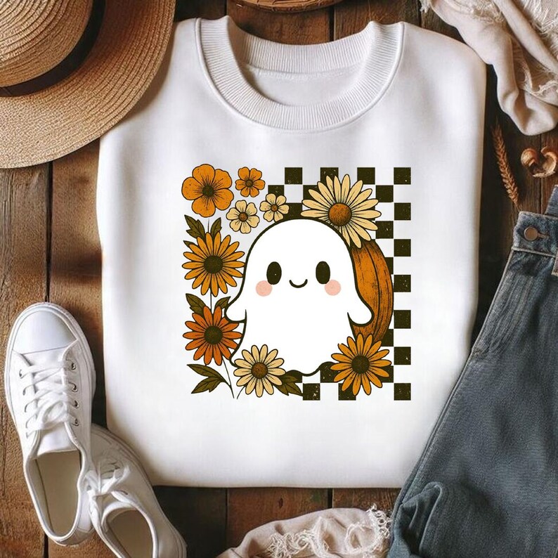 Pode incluir: Uma camisola branca de gola redonda com um gr&aacute;fico central de um fantasma branco sorridente com bochechas cor-de-rosa. O fantasma est&aacute; rodeado por flores cor de laranja e amarelas, uma ab&oacute;bora e um padr&atilde;o axadrezado preto e branco.