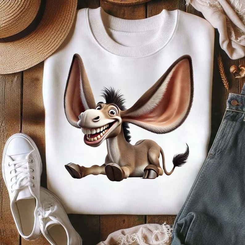 Op de afbeelding: Witte sweatshirt met een cartoon ezel met grote oren en een brede grijns. De ezel is bruin en bruin met een zwarte manen en staart. Het sweatshirt ligt op een houten oppervlak met een strohoed, witte sneakers en een spijkerbroek.