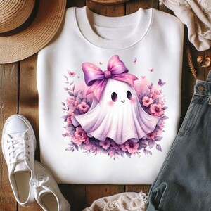 Pode incluir: Uma camisola branca de gola redonda com um gr&aacute;fico central de um pequeno fantasma branco com um grande la&ccedil;o cor-de-rosa na cabe&ccedil;a. O fantasma est&aacute; rodeado por flores cor-de-rosa e roxas e pequenas borboletas, criando uma ilustra&ccedil;&atilde;o suave e colorida.