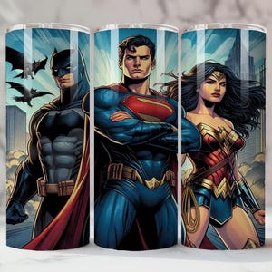 May include: Three stainless steel tumblers featuring DC Comics superheroes Batman, Superman, and Wonder Woman. The tumblers have a blue background with cityscapes and illustrations of the characters in their iconic costumes. The Batman tumbler has bats flying in the background.