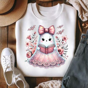 Pode incluir: Uma camisola branca de gola redonda com um gr&aacute;fico central de um fantasma de desenho animado com um la&ccedil;o rosa brilhante, a ler um livro rosa. O fantasma est&aacute; rodeado por flores cor-de-rosa e pequenos morcegos, com uma bainha de renda.