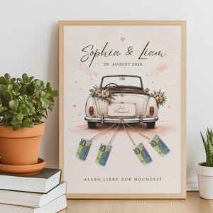 Op de afbeelding: Een ingelijste print met een vintage cabriolet, bloemstukken, een Just Married-bord en vier opgerolde biljetten van twintig euro bevestigd met touwtjes. De tekst luidt Sophia & Liam, 20. AUGUST 2026 en ALLES LIEBE ZUR HOCHZEIT.