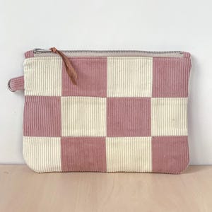 Könnte beinhalten: Eine rosafarbene und cremefarbene Cord-Reißverschlusstasche mit Schachbrettmuster. Die Tasche hat eine kleine Schlaufe an der Seite und einen Leder-Reißverschlussanhänger. Hintergrund: helle Holzoberfläche und weiße Wand.