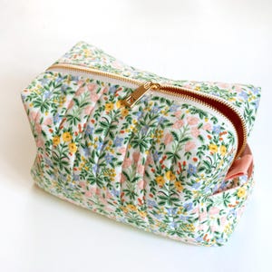 Puede incluir: Un neceser con estampado floral y cremallera dorada. La bolsa presenta un fondo blanco con un estampado de flores rosas, amarillas y azules y hojas verdes. El forro interior es de color melocotón liso.