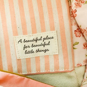 Op de afbeelding: Een stoffen etui met een cr&egrave;me en perzikkleurig gestreept patroon en een rechthoekig label op de voorzijde. Het label bevat de zwarte tekst A beautiful place for beautiful little things. Een bloemenstof met roze bloemen is zichtbaar.