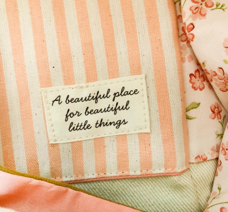 Op de afbeelding: Een stoffen etui met een cr&egrave;me en perzikkleurig gestreept patroon en een rechthoekig label op de voorzijde. Het label bevat de zwarte tekst A beautiful place for beautiful little things. Een bloemenstof met roze bloemen is zichtbaar.