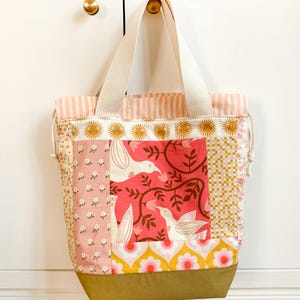 Op de afbeelding: Een patchwork draagtas met diverse stofpatronen, waaronder roze bloemen, een centraal paneel met witte vogels op een rode achtergrond, gele en roze bloemmotieven en een effen olijfgroene onderkant met cr&egrave;mekleurige canvas hengsels.