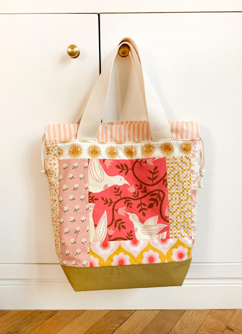 Op de afbeelding: Een patchwork draagtas met diverse stofpatronen, waaronder roze bloemen, een centraal paneel met witte vogels op een rode achtergrond, gele en roze bloemmotieven en een effen olijfgroene onderkant met cr&egrave;mekleurige canvas hengsels.