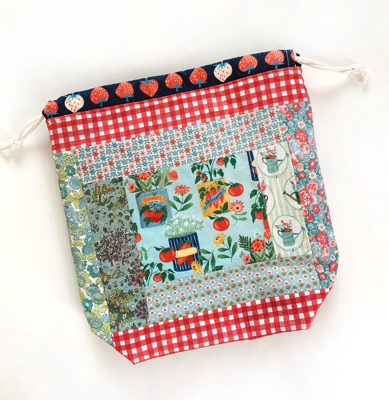 May include: A fabric drawstring bag featuring a patchwork design with various patterns including red and white gingham, a light blue panel with tomato and garden illustrations, floral prints, and a dark blue top border with a strawberry pattern.
