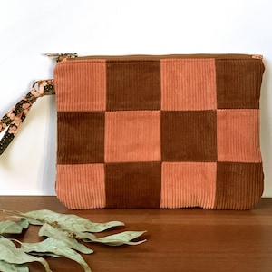 May include: A corduroy clutch with a checkerboard pattern in shades of brown and peach. The clutch has a wrist strap with a floral design and a zipper closure. The bag is rectangular and sits on a wooden surface.