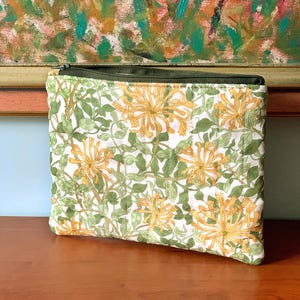 May include: A rectangular zipped pouch with a floral design. The pouch has a white background with yellow flowers and green leaves. The zip is dark green, and the pouch is on a wooden surface.