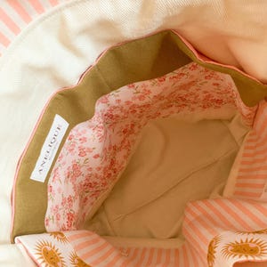Op de afbeelding: De binnenkant van een stoffen tas met een cr&egrave;mekleurige basis en een roze-wit gestreepte rand. De voering heeft een bloemenpatroon met kleine roze bloemetjes en een sectie met een zonnetjesprint. Een wit label bevat de tekst ANELIQUE.