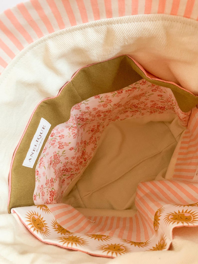 Op de afbeelding: De binnenkant van een stoffen tas met een cr&egrave;mekleurige basis en een roze-wit gestreepte rand. De voering heeft een bloemenpatroon met kleine roze bloemetjes en een sectie met een zonnetjesprint. Een wit label bevat de tekst ANELIQUE.