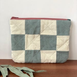 May include: A quilted corduroy pouch with a checkerboard pattern in light blue and cream. The pouch has a burgundy zipper and a rectangular shape. The fabric has a ribbed texture.