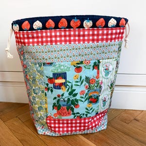 May include: A fabric drawstring bag featuring a top band with a strawberry print on a dark blue background, a red and white gingham section, and a central panel displaying illustrations of tomato plants in cans and various garden flowers.