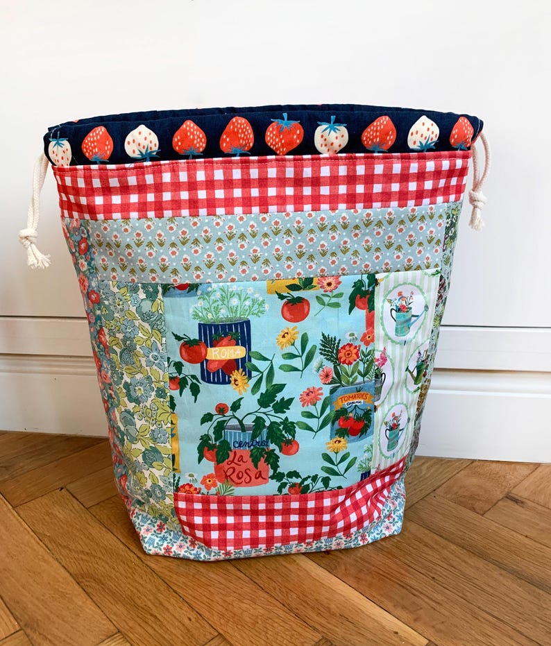 May include: A fabric drawstring bag featuring a top band with a strawberry print on a dark blue background, a red and white gingham section, and a central panel displaying illustrations of tomato plants in cans and various garden flowers.