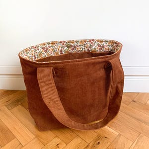 Könnte beinhalten: Eine rostfarbene Cord-Tragetasche mit zwei passenden Schulterriemen. Das Innenfutter zeigt ein dichtes Blumenmuster mit kleinen Blüten in Orange-, Gelb-, Rosa- und Grüntönen. Ein einzelner Metalldruckknopf ist am oberen Rand der Tasche zu sehen.