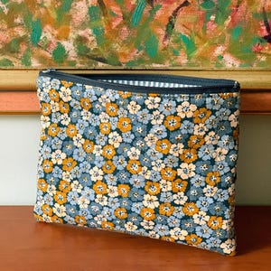 Op de afbeelding: Een rechthoekig etui met bloemenprint en een donkerblauwe rits. De etui heeft een patroon van kleine blauwe, gele en witte bloemen op een donkerblauwe achtergrond. De binnenvoering is een wit en blauw gestreepte stof.