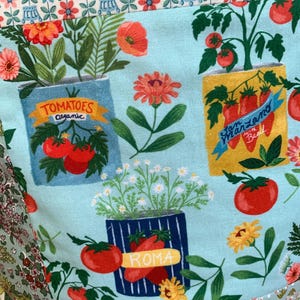May include: A light blue fabric featuring a repeating pattern of potted tomato plants in various containers labelled Tomatoes Organic, San Marzano with Basil, and Roma, interspersed with orange and pink flowers and green foliage.